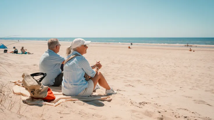 Ouder stel zit op een strandlaken met tas en eten aan het strand