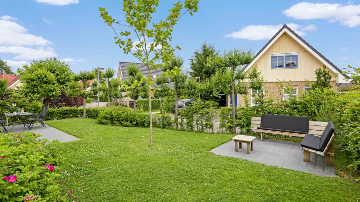 Tuin met grasveld, loungebank en jonge boom naast een vakantiewoning op het park
